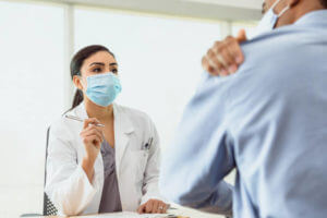 A mature man rubs his shoulder as he tells his doctor about his shoulder pain during a medical appointment. The doctor listens attentively to the patient.